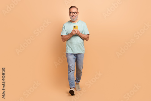 Full size portrait of positive cheerful aged person hold smart phone walking isolated on beige color background