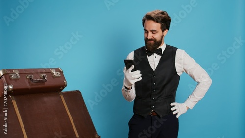 Cheerful employee browses mobile phone, feeling joyful about positive news on online webpages over background. Happy remarkable doorman with white gloves being excited on camera.