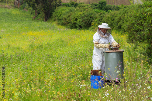 mujer latina en medio de un campo verde con una maquina centrifugadora manual sacando miel de abeja de unos panales llenas de miel la apicultora esta vistiendo un traje de seguridad de color blanco