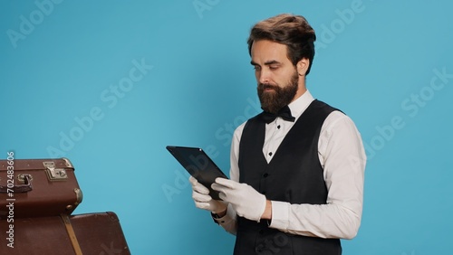 Male bellhop working on tablet, being in charge of trolley bags for safekeeping. Doorman hotel porter presents commitment for welcoming atmosphere, symbolizing his role in hospitality industry.