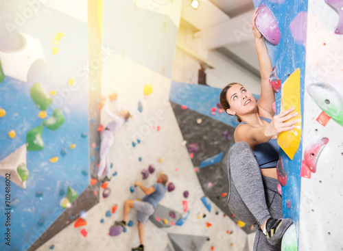 Concentrated young girl in comfortable athletic clothing moving carefully on bouldering wall clinging to colorful hand and foot holds. Popular activity at indoor climbing gym.