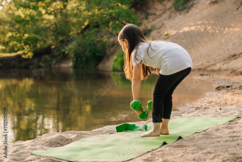 Junior female athlete bent down to put small green dumbbells on sporty mat. Little girl training on beach near river. photo