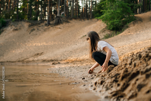 Canvas Print Curious girl with brown hair squatting down on sand near river in forest and going to take grit