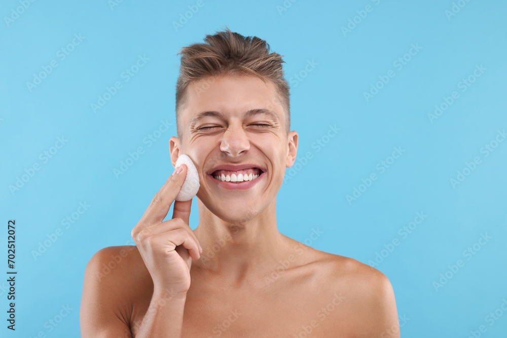 Happy young man washing his face with sponge on light blue background Happy young man washing his face with sponge on light blue background