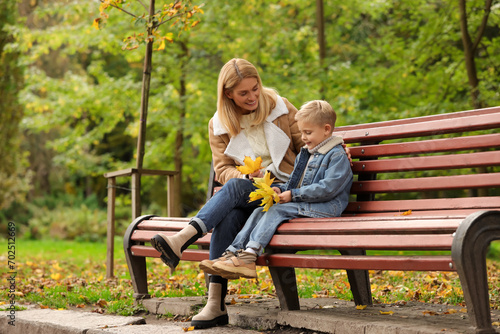 Happy mother and her son spending time together with dry leaves on wooden bench in autumn park