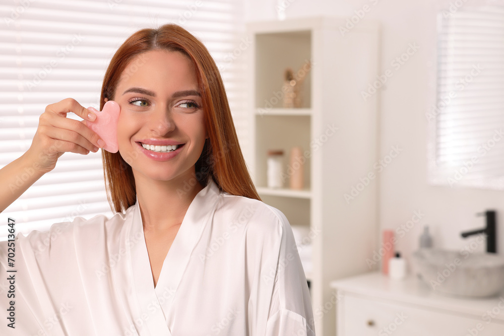 Young woman massaging her face with rose quartz gua sha tool in bathroom, space for text