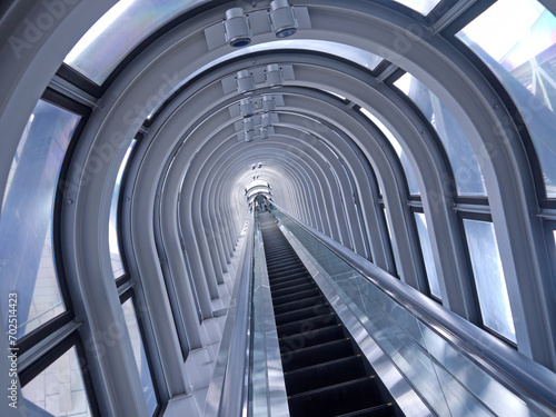 Going up on Long straight escalator in glass tunnel with steel arches, perspective view