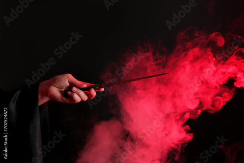 Magician holding wand in smoke on dark background, closeup photo