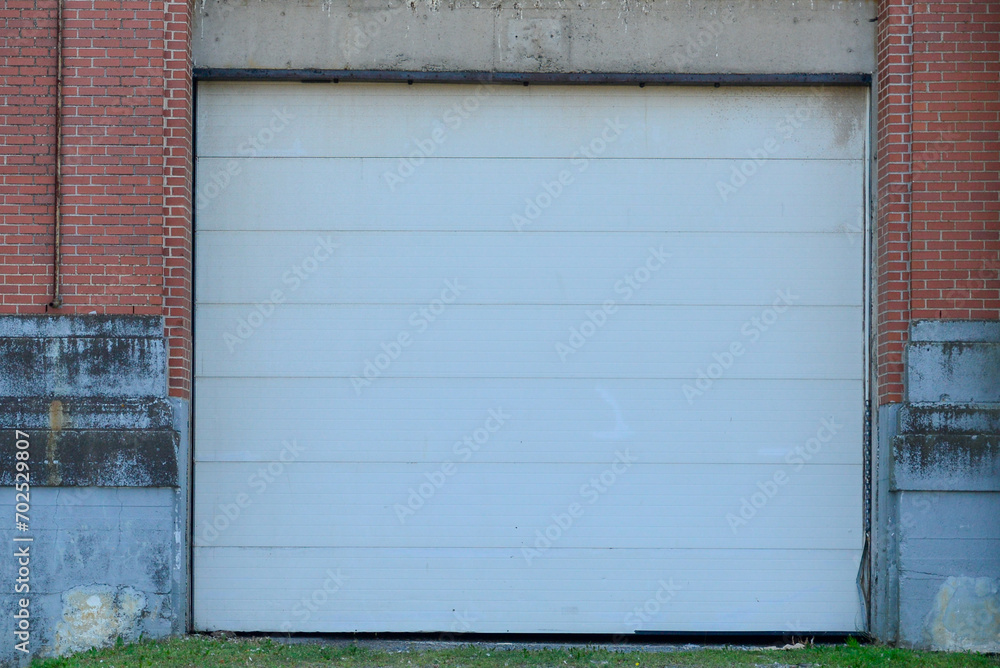 A large white metal panel car garage door in a narrow red brick building. The foundation of the building is grey concrete with a black waterproof coating. The ground in front is asphalt. A large white metal panel car garage door in a narrow red brick building. The foundation of the building is grey concrete with a black waterproof coating. The ground in front is asphalt.