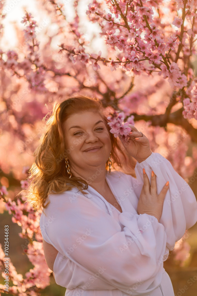 Fototapeta premium Woman blooming peach orchard. Against the backdrop of a picturesque peach orchard, a woman in a long white dress enjoys a peaceful walk in the park, surrounded by the beauty of nature.