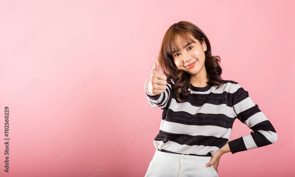 A smiling Asian woman in a studio shot on pink background confidently gives a thumbs-up 'Ok' sign, symbolizing agreement and success. Her cheerful expression radiates positivity and satisfaction. A smiling Asian woman in a studio shot on pink background confidently gives a thumbs-up 'Ok' sign, symbolizing agreement and success. Her cheerful expression radiates positivity and satisfaction.