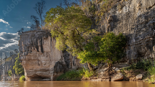 The steep banks of the river. Trees grow on limestone cliffs. Clouds in the blue sky. Highlights on the red-brown water. Madagascar. Manambolo river.