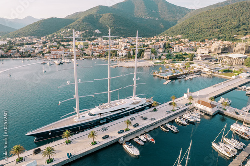 Long sailing yacht is moored at the pier against the background of the coast with modern hotels and high-rise buildings. Porto, Montenegro. Drone