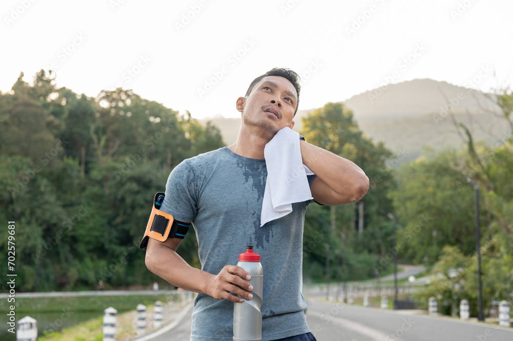 Fototapeta premium A tired Asian man is wiping his sweat on his face, resting after a long run outdoors in the morning.