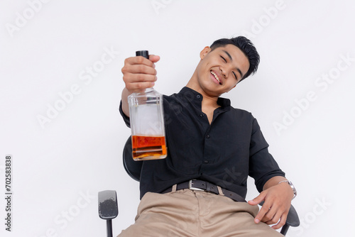 A visible tipsy but friendly young man offers a half-empty bottle of whiskey while slouching on the chair. Isolated on a white backdrop.