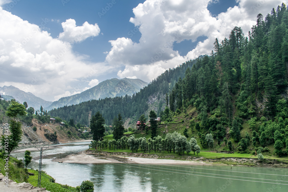 Teal colored River Neelum on a cloudy day with beautiful forest, green meadow and white sand on its banks