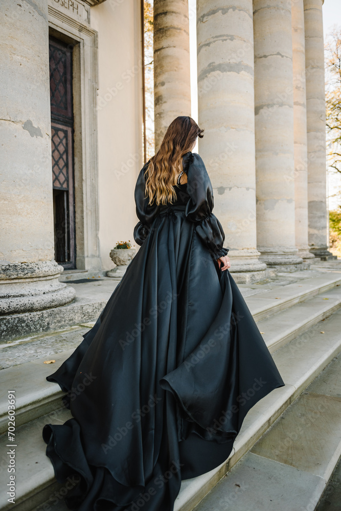 Naklejka premium Bride in black dress walks on stairs near large columns of ancient temple at sunset. Female near ancient Baroque Roman Catholic church in Pidhirtsi, Lviv, Ukraine. Girl in nature autumn day. Back view