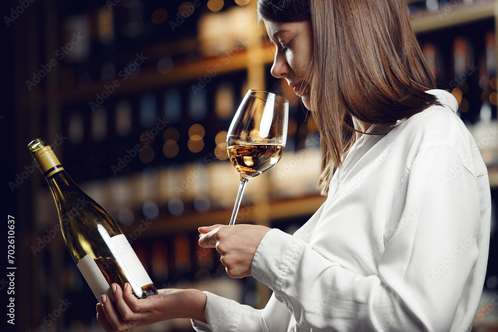 Focused female wine expert smells wine into glass, holding bottle and reads label with characteristics of wine, the year of manufacture, region and the degree of acidity of the alcoholic beverage. - obrazy, fototapety, plakaty 