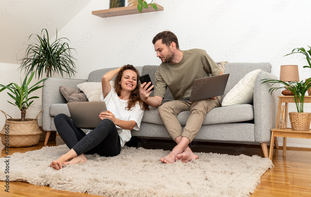 Young couple at home uses laptops and mobile phone, man showing screen of phone to woman. Young couple at home uses laptops and mobile phone, man showing screen of phone to woman.