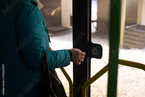 Elderly woman in a warm jacket with a bag in her hand reaches for the button to open the glass doors from inside a modern city bus
