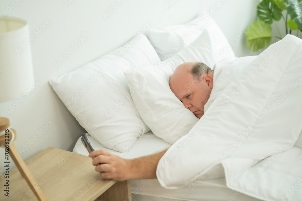 Young over weight man in bed, waking up, looking at alarm clock at smartphone with surprising time. Lazy day in morning on bed. Young over weight man in bed, waking up, looking at alarm clock at smartphone with surprising time. Lazy day in morning on bed.