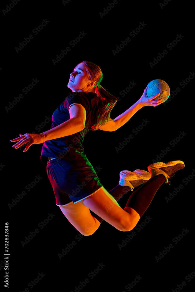 Action shot of young woman, professional handball player passionately playing handball, executing powerful throw against black background in neon light. Concept of sport, energy, dynamic, championship