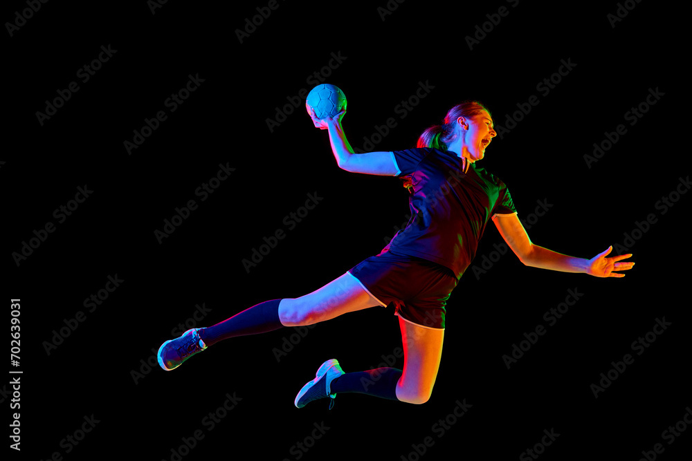 Fit, focused woman engaged in handball drills, displaying determination and focus against black background in mixed neon light. Concept of sport, hobby, movement, dynamic, championship, goal. Ad