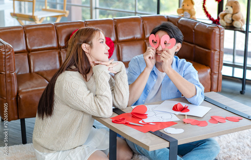 Asian young handsome male boyfriend sitting on sofa smiling cuddling with beautiful female girlfriend holding red heart shape paper cutting in romantic decorated living room celebrating valentine day photo