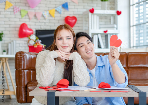 Asian young handsome male boyfriend sitting on sofa smiling cuddling with beautiful female girlfriend holding red heart shape paper cutting in romantic decorated living room celebrating valentine day photo