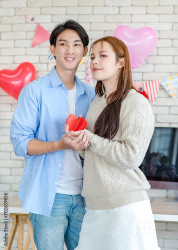 Asian young handsome male boyfriend standing smiling playing cuddling with beautiful female girlfriend holding red heart shape paper cutting in romantic decorate living room celebrating valentine day photo