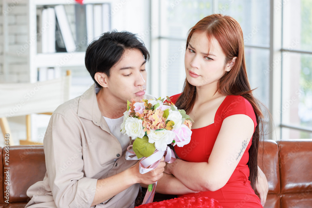 Millennial Asian young handsome male boyfriend reconciling beautiful female sulking upset angry girlfriend in red dress with white roses flower bouquet sit crossed arms on sofa in living room at home