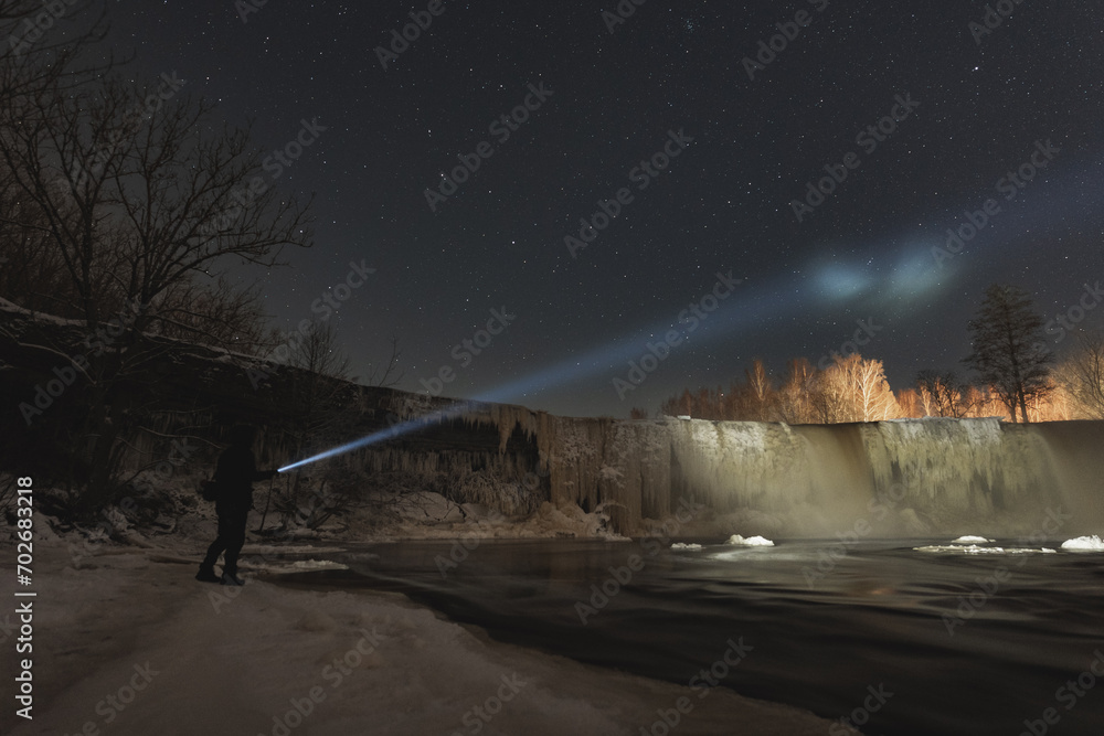 Man with a flashlight at Jagala waterfall in winter at night.