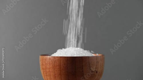 White table salt granules in a pile with spoon. Wood bowl. Gray color studio background. Condiment particles spinning. Clean cosmetic bath salt footage. Closeup of grained sea salt pieces hill	 photo