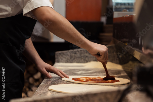 A chef in a professional kitchen applies tomato sauce evenly on stretched pizza dough, showcasing pizza preparation