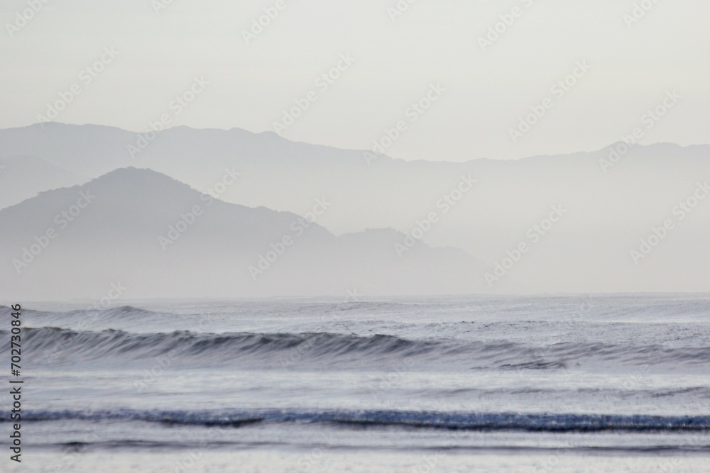 Blurred seascape with foggy morning on seashore. Light blue sea, small waves, mountains silhouette, cloudy sky.  