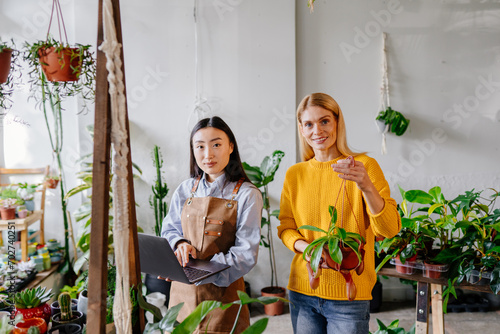 Happy middle age woman in yellow sweater in a flower shop with a purchased plant in her hands standing with asian female seller in apron holding laptop surround plants in plant store interior.