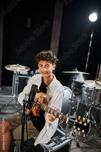 cheerful talented teenage boy in casual outfit playing his guitar and looking at camera in studio