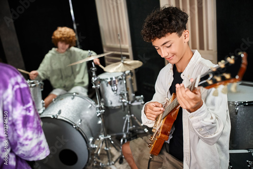 Fotografi focus on cheerful cute teenage boy playing guitar actively next to his blurred b