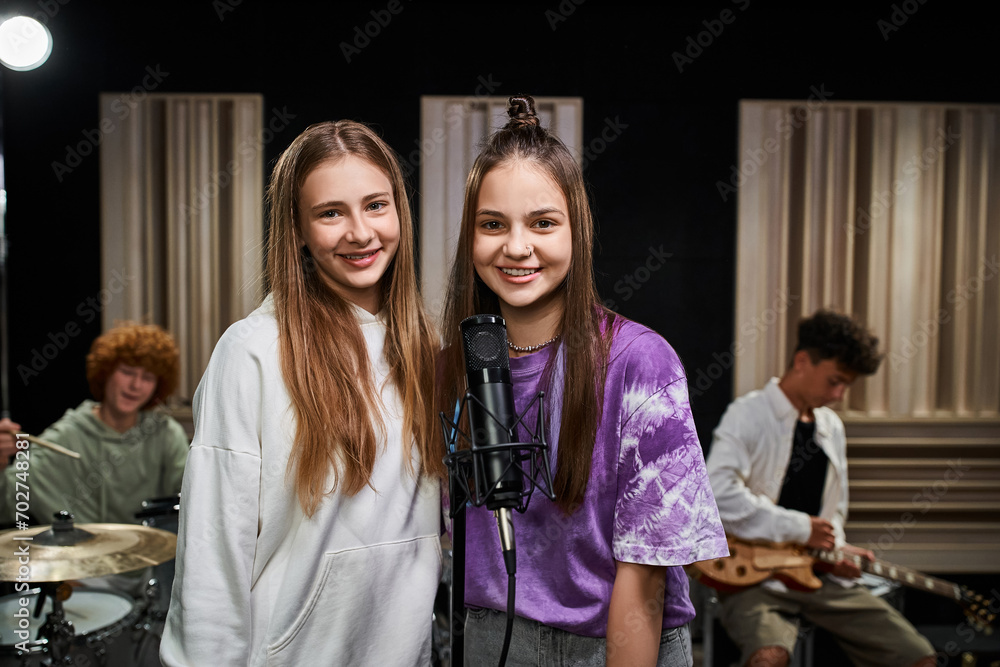 jolly teenage girls looking at camera near microphone while their friends playing drums and guitar jolly teenage girls looking at camera near microphone while their friends playing drums and guitar