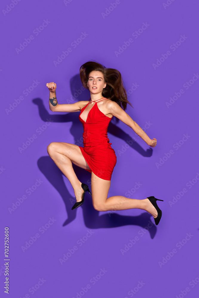 Charming young woman wearing in fashion red dress posing, running in motion in mid-air against purple background. Concept of fashion and beauty, modern style, shopping, sales season. Ad