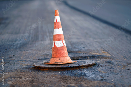 Orange traffic cone stands on manhole. Road repair works, asphalt paving, pylon to mark an obstacle or hole on road. Traffic cone stands on hatch. Road reconstruction, renew old asphalt surface photo