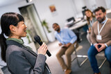 Young business woman lecturing her multiethnic colleagues. Having a speech while holding a microphone.