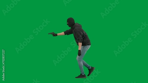 Portrait of thief on chroma key green screen background. Man robber in balaclava and hoodie walking holding gun in hand looks around. Side view.