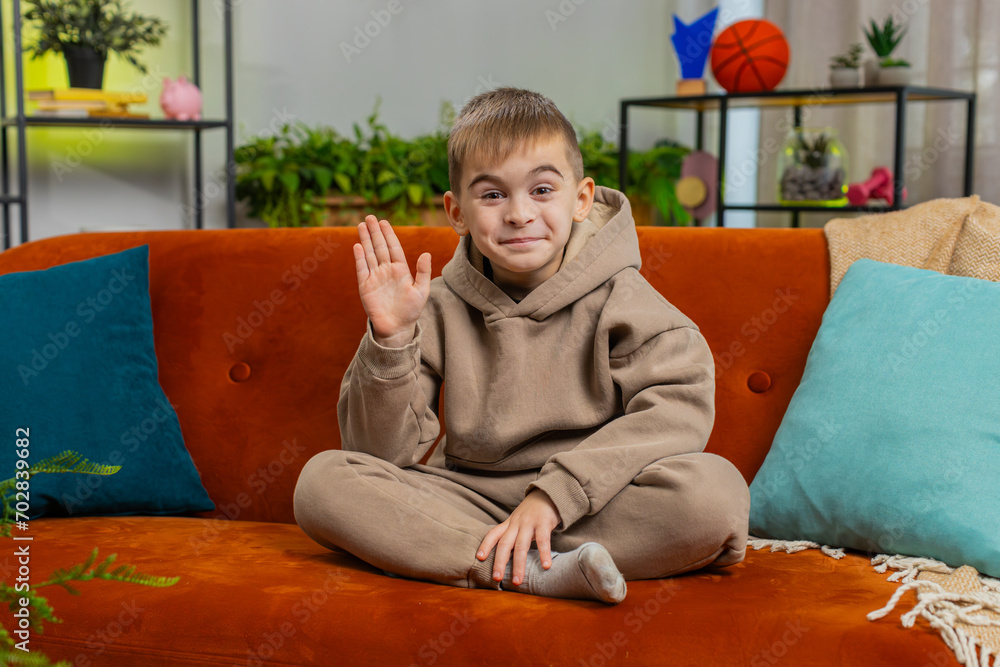 Obraz premium Young child boy smiling friendly at camera and waving hands gesturing hello, hi, greeting or goodbye, welcoming with hospitable expression. Teenager Caucasian kid at home in living room sits on sofa