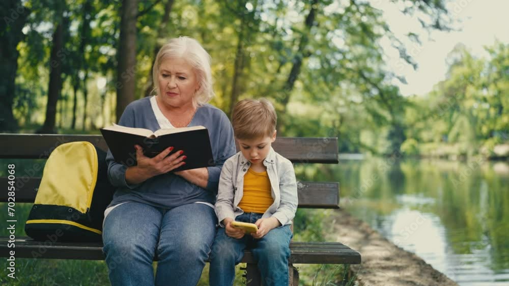 Senior lady reading book, little boy playing on gadget in park, generation gap Senior lady reading book, little boy playing on gadget in park, generation gap