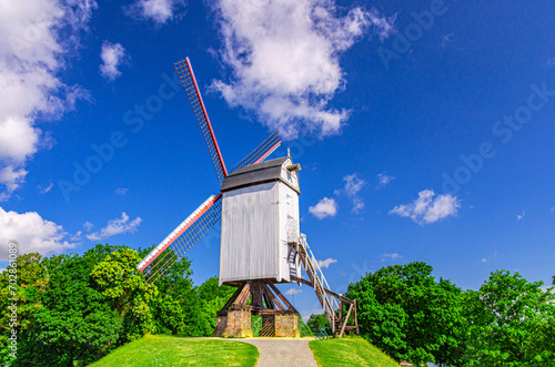 Traditional old windmill on green hill in Brugge city, blue sky white clouds background in summer sunny day, white wooden wind mill with sails, path in park with green trees, Flemish Region, Belgium