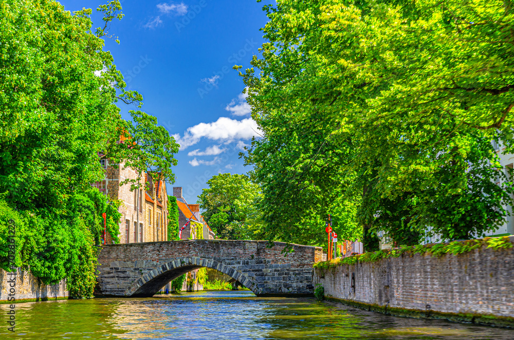 Meebrug stone bridge across Groenerei Green Canal water of Reie river, green trees on embankment, Brugge old town, medieval buildings in Bruges city historical center, Flemish Region, Belgium Meebrug stone bridge across Groenerei Green Canal water of Reie river, green trees on embankment, Brugge old town, medieval buildings in Bruges city historical center, Flemish Region, Belgium