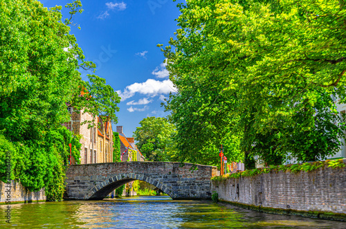 Meebrug stone bridge across Groenerei Green Canal water of Reie river, green trees on embankment, Brugge old town, medieval buildings in Bruges city historical center, Flemish Region, Belgium