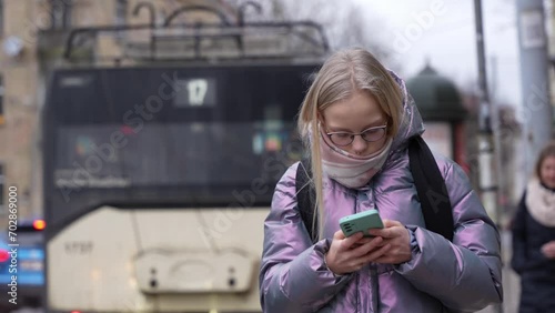 Close-up camera view of teen girl standing in the bus stop and scrolling her phone. Young girl waits for bus to school. Schoolgirl with glasses and backpack. photo