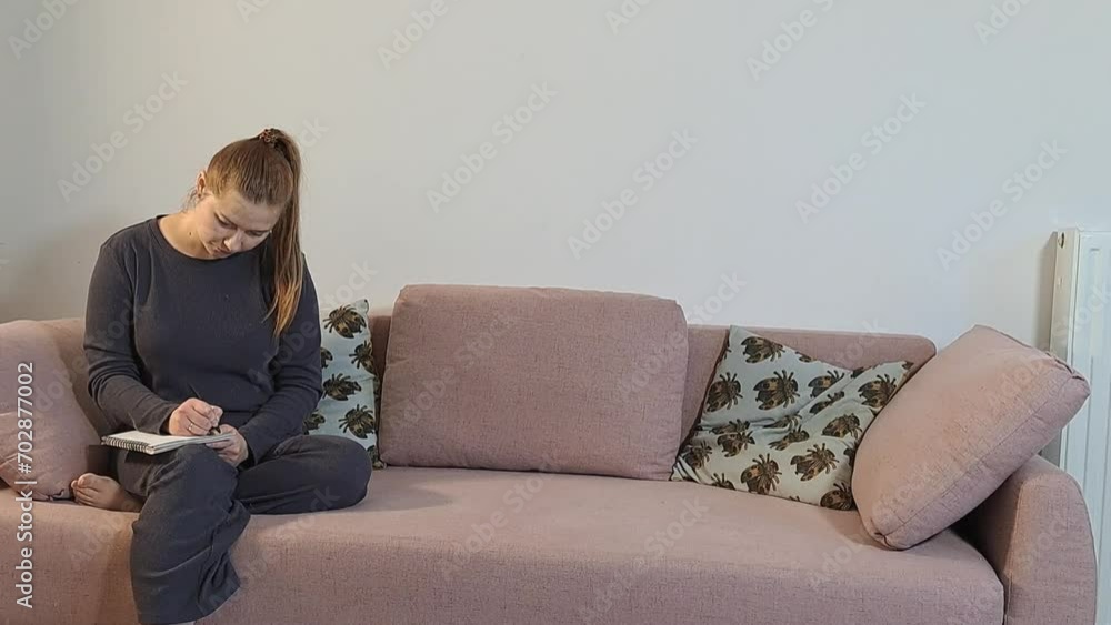 Young red woman writing on notebook sitting on couch at cozy home. Working or studying and thinking at home office Young red woman writing on notebook sitting on couch at cozy home. Working or studying and thinking at home office